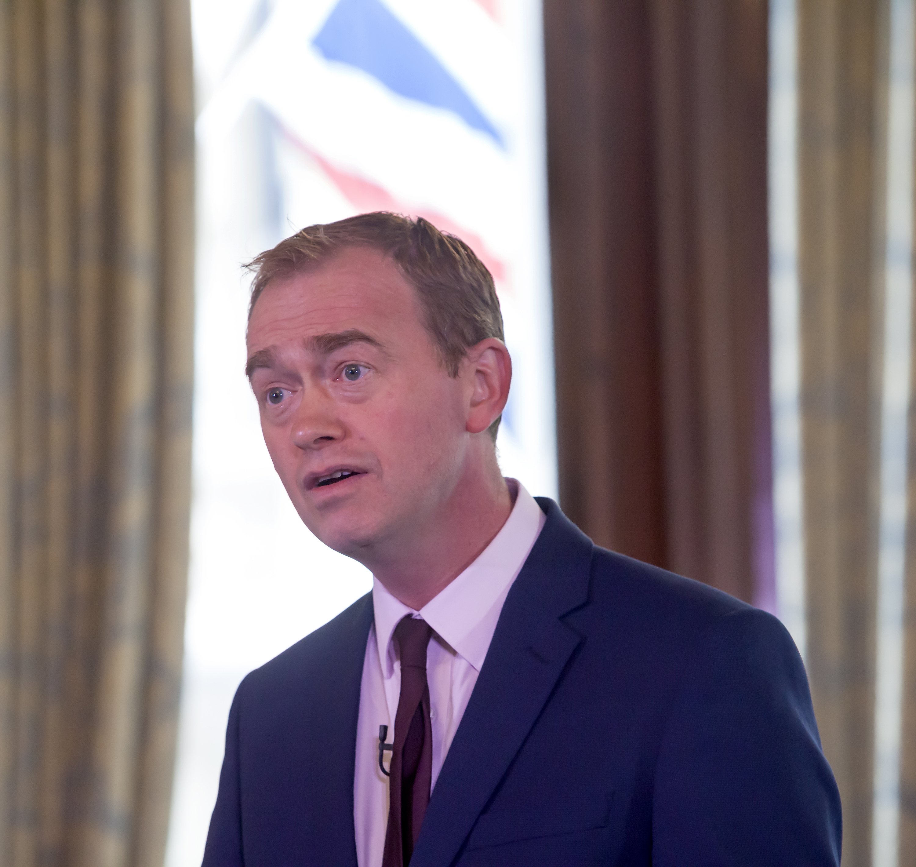 Politician answers questions in a press conference, wearing a microphone and with a backdrop of curtains and closed window