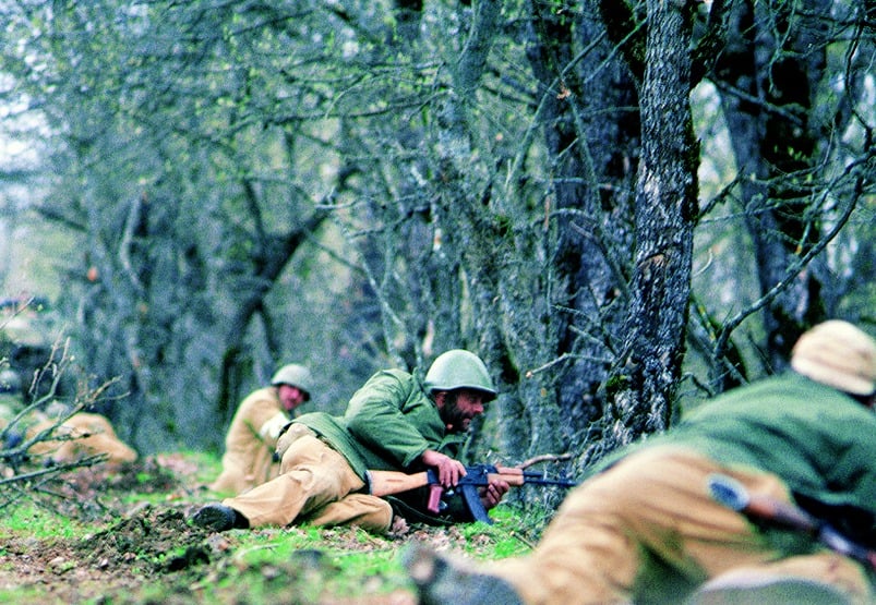 Soldiers in green uniforms and helmets lie prone in a forest, aiming rifles. Sparse trees and green undergrowth surround them.