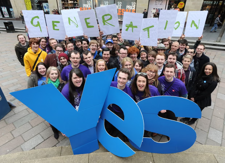 A group of smiling people in purple shirts holding a large blue "Yes" sign and letters spelling "Generation" on signs, standing in a shopping street.