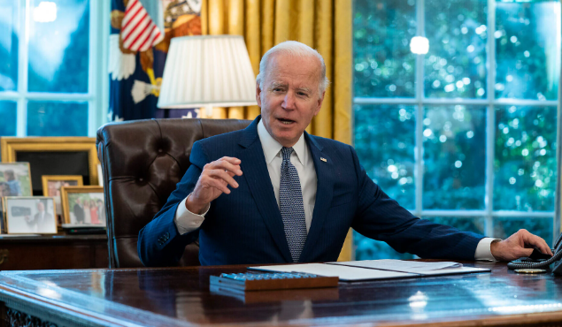 Man in a blue suit sits at an office desk, speaking and gesturing with his hand. Behind him are gold curtains and a window with green foliage visible.