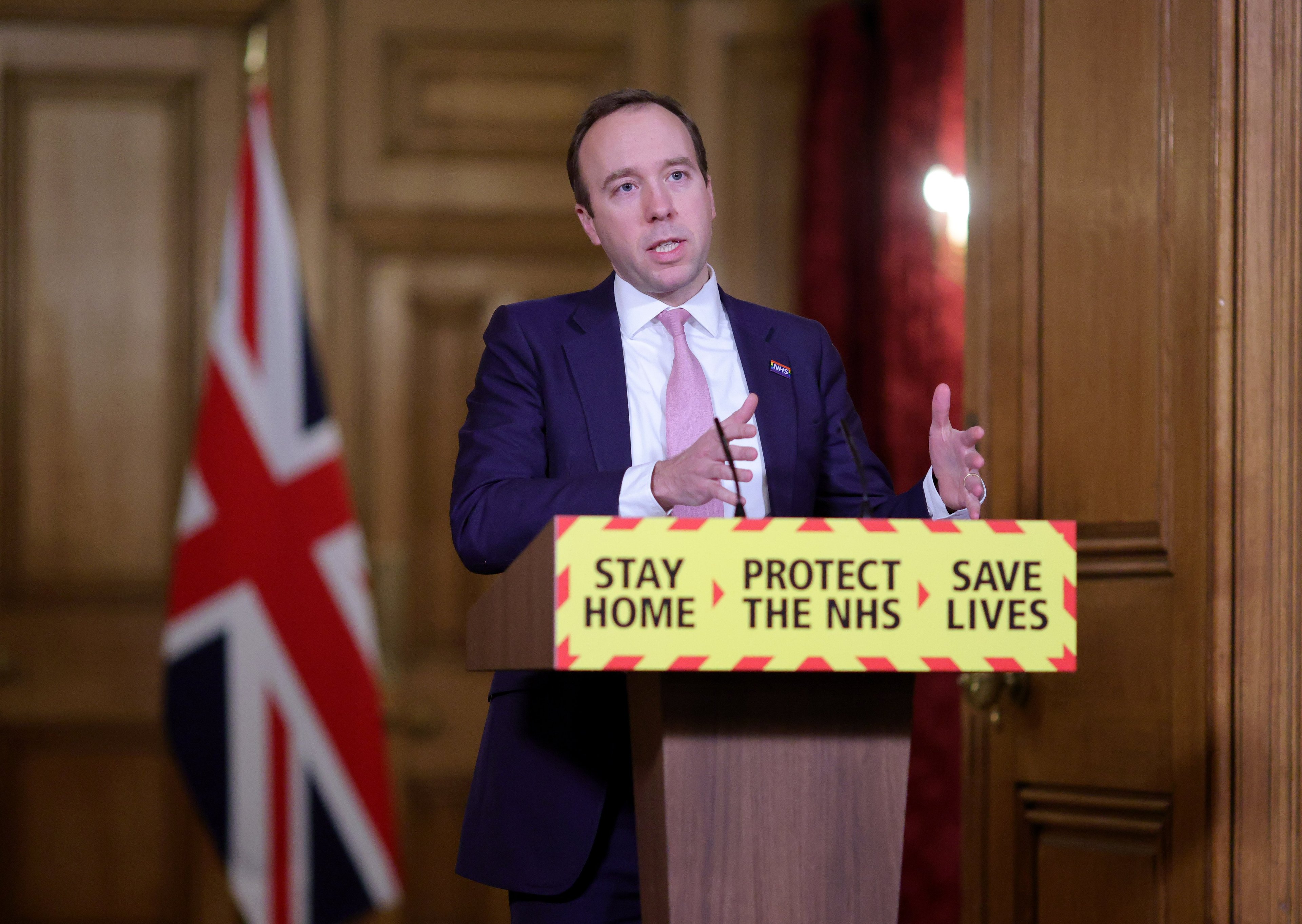 A man stands at a lectern, speaking to the press with union flags in the background the the slogan 'Stay Home, Protect the NHS, Save Lives' in the foreground