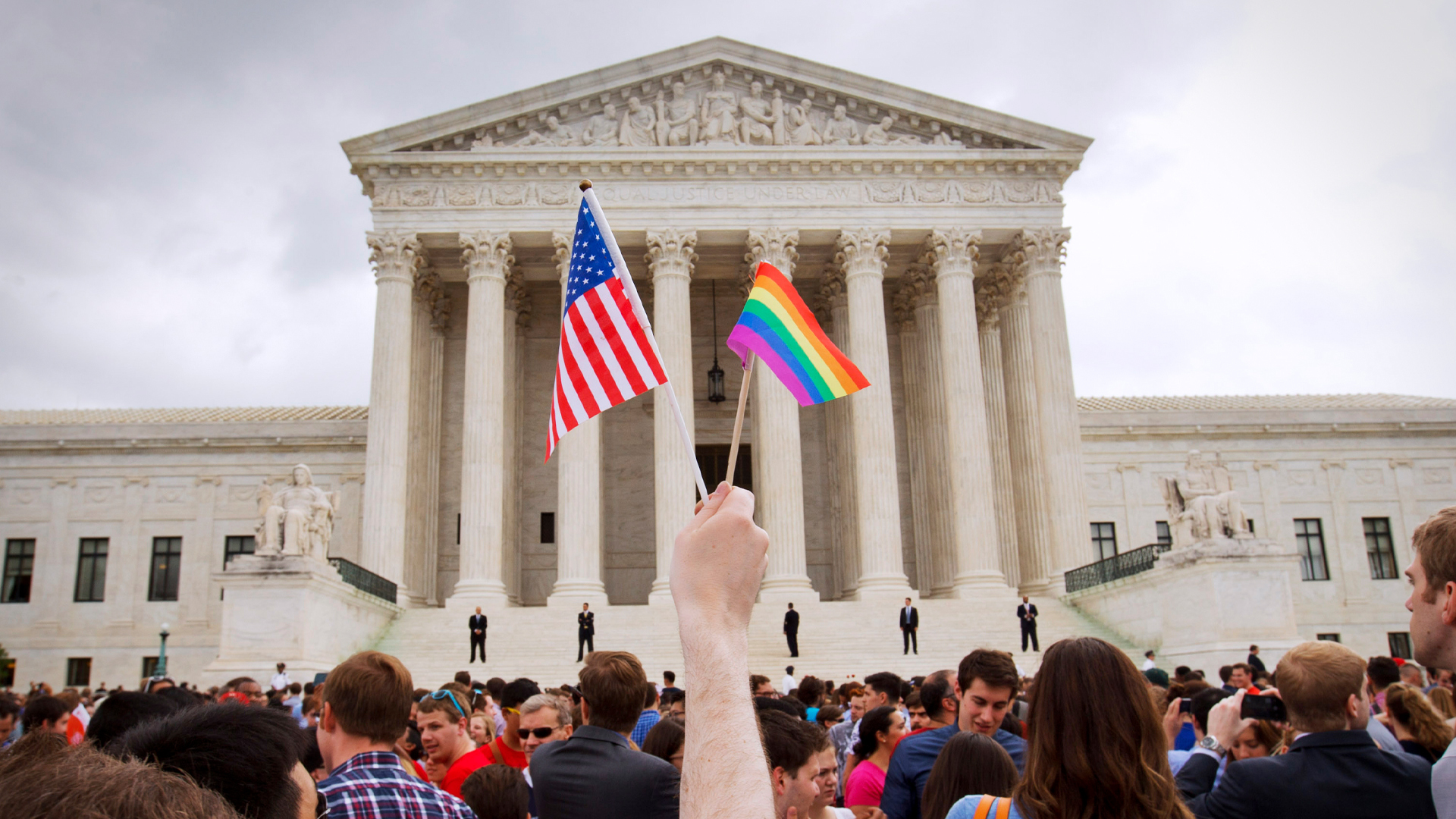 Crowd outside a government building with a hand holding two flags, the American flag and a rainbow flag, symbolising pride and unity.
