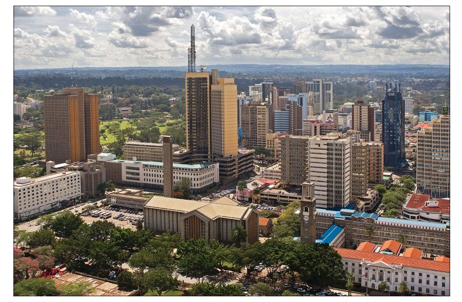 Skyline of a city with modern skyscrapers and historic buildings, surrounded by green trees under a partly cloudy sky.
