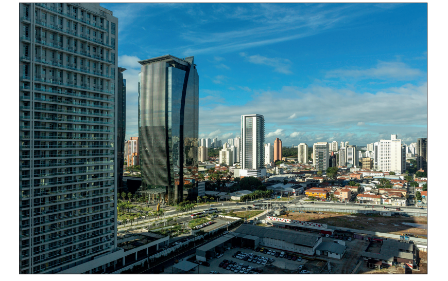 Cityscape with modern skyscrapers in the foreground and smaller buildings in the background, under a blue sky with light clouds.