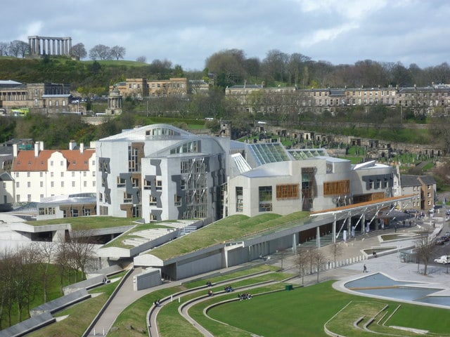 Modern building with unique architecture, green roofs, open courtyard, and surrounding parkland. Historical structures and hills are visible in the background.