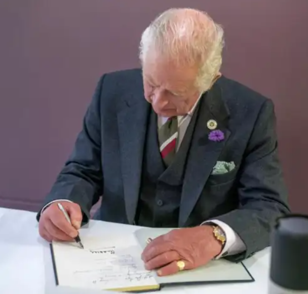 Elderly man in a suit and tie signing a document at a table, wearing a gold watch and a purple flower pinned to his lapel.