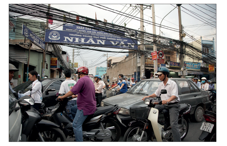 Busy urban street scene with numerous motorbikes and people navigating traffic near a school gate under power lines and signage.