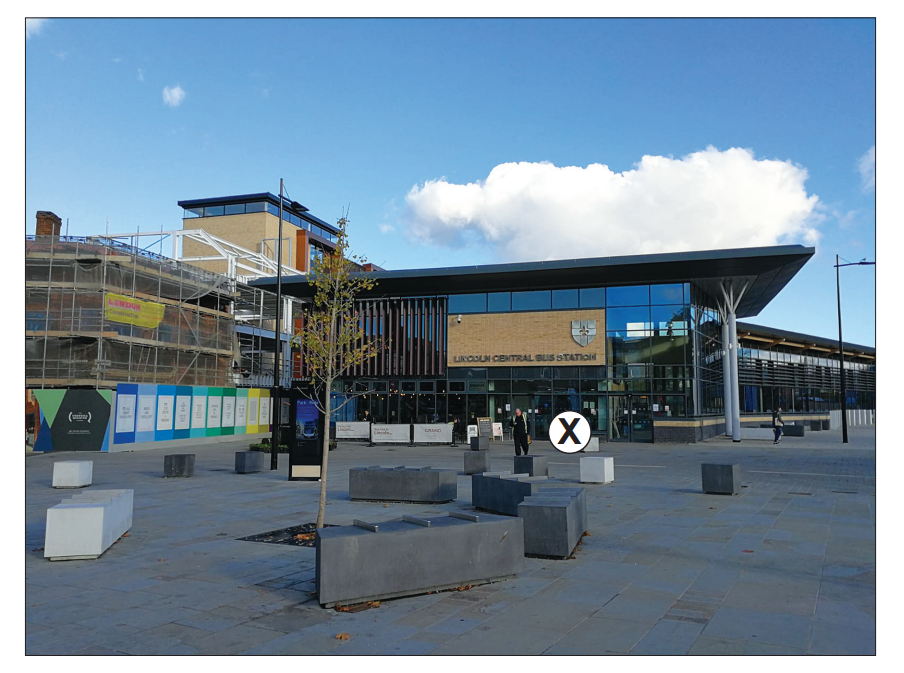 Modern bus station with glass facade, surrounded by a paved plaza and seating, construction site on the left, under a partly cloudy sky.