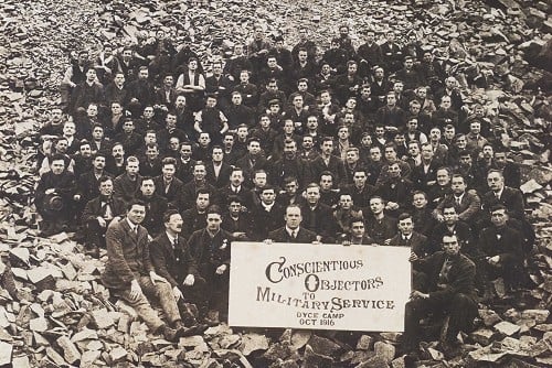 A large group of men, likely early 20th century, pose on rocky ground holding a sign reading "Conscientious Objectors to Military Service, Dyce Camp, Oct 1916".