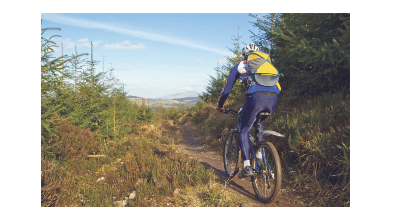 Person cycling on a dirt path through a forested area with distant hills under a clear sky, wearing a helmet and carrying a yellow backpack.