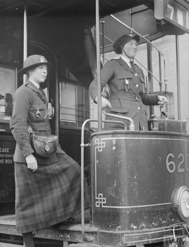 Two women in early 20th-century uniforms operate a vintage tram, one standing and the other seated, both wearing hats, beside tram number 620.