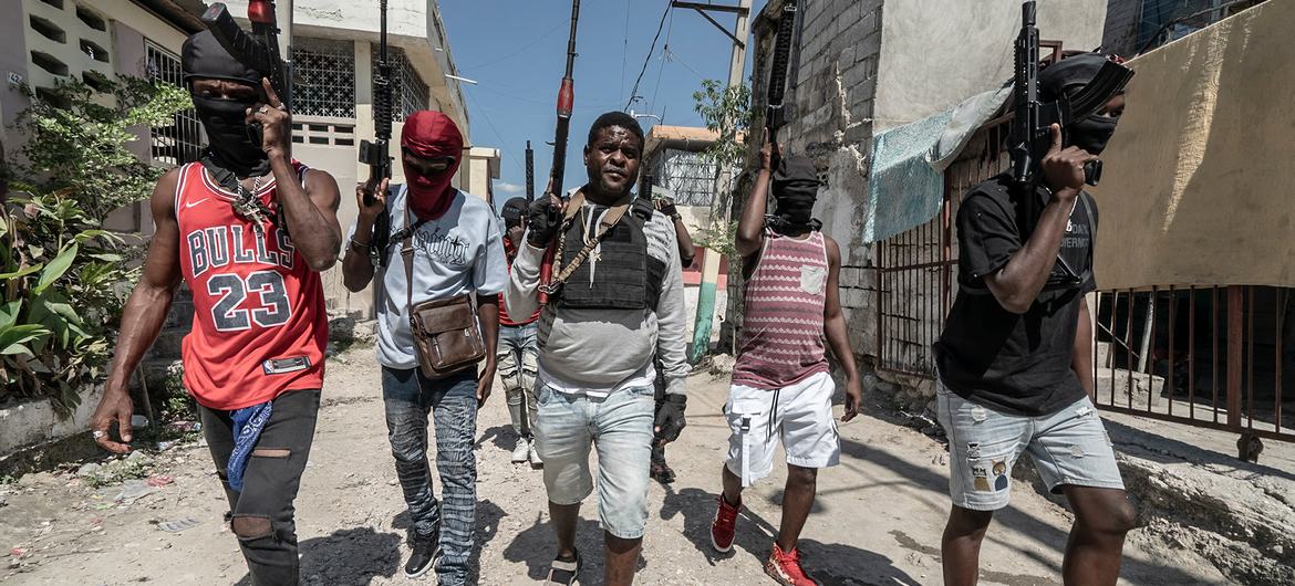 Group of masked men with firearms walk down a narrow street in a residential area, with a mix of concrete and makeshift buildings in the background.