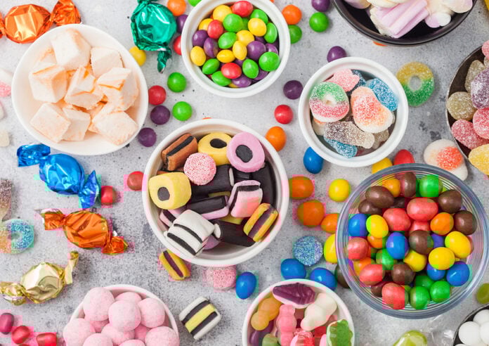 Various colourful sweets in bowls on a grey background, including liquorice, marshmallows, gummy candies, and chocolate-coated pieces.