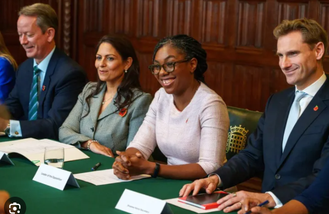 A group of four people seated at a green table with nameplates, dressed in business attire, in a wood-panelled room with ornate carvings.