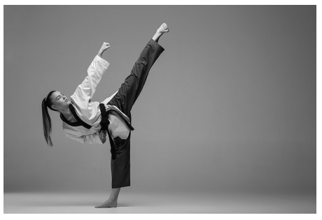 Martial artist performing a high kick, wearing a traditional uniform with a black belt, in a minimal studio setting, against a plain grey background.