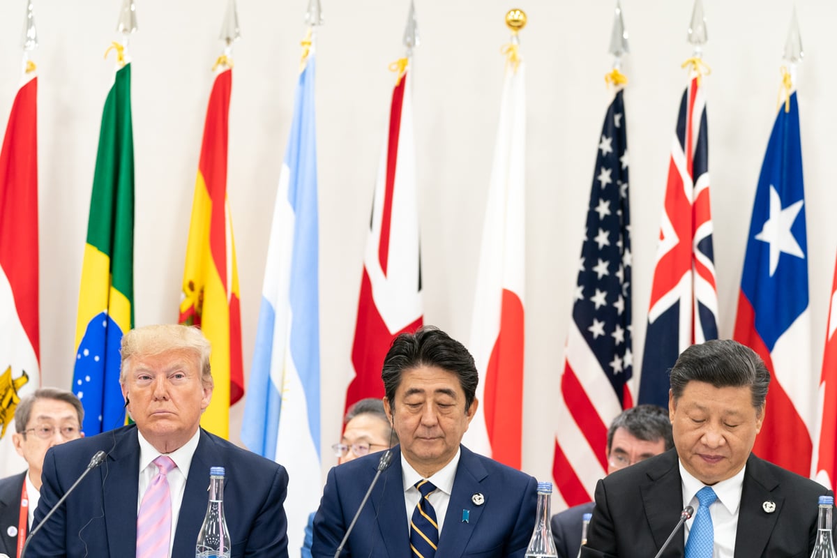 Three men in suits sit at a table with microphones, in front of multiple international flags; the central man has his eyes closed.
