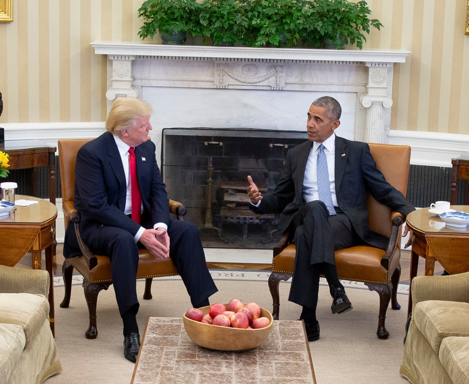 Two men in suits are sitting in armchairs in an office with a fireplace and plants. A bowl of apples is on the table between them.