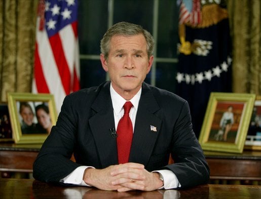 Man in a suit and red tie sits at a desk with hands clasped, framed photos and US flags in the background, suggesting a formal setting.