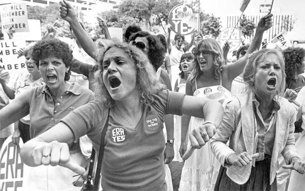 Women passionately protest for equal rights, wearing ERA shirts and holding signs. The crowd shows determination and solidarity in an outdoor setting.