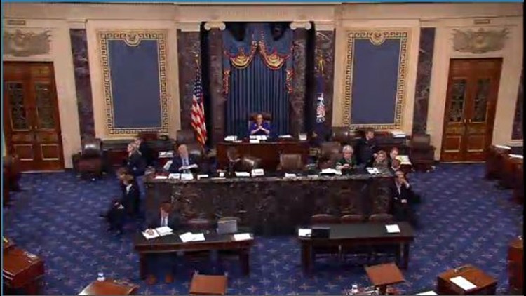 People seated at desks in a formal government chamber with ornate blue walls and flags, suggesting a legislative or parliamentary setting.