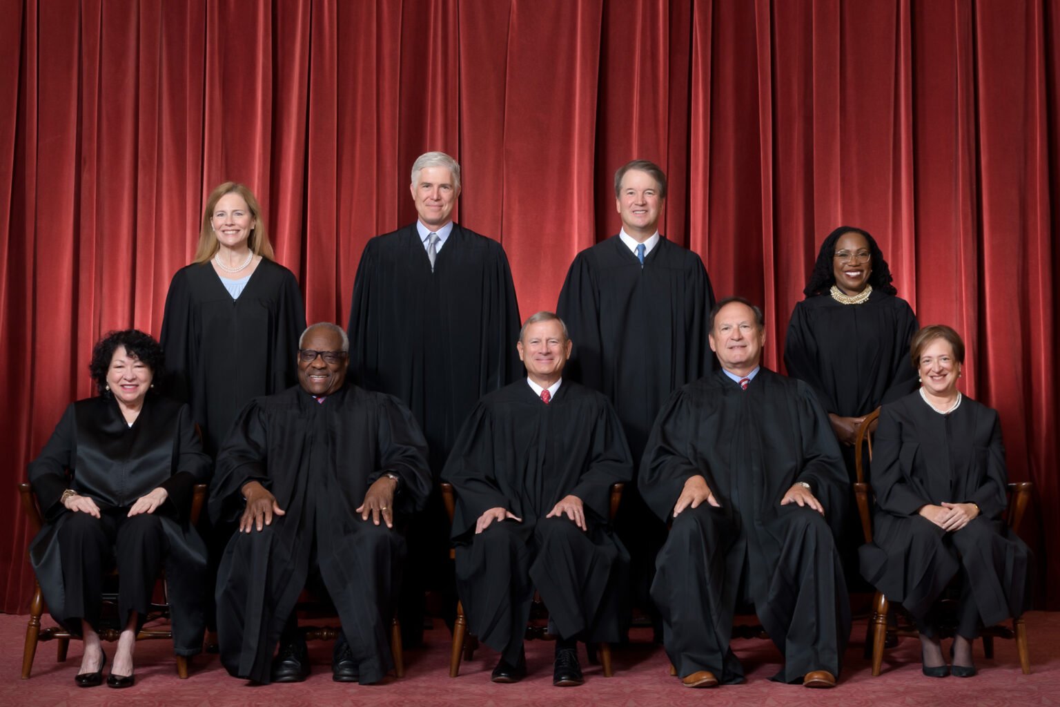Nine judges in black robes pose in two rows against red curtains; five seated in front and four standing behind.
