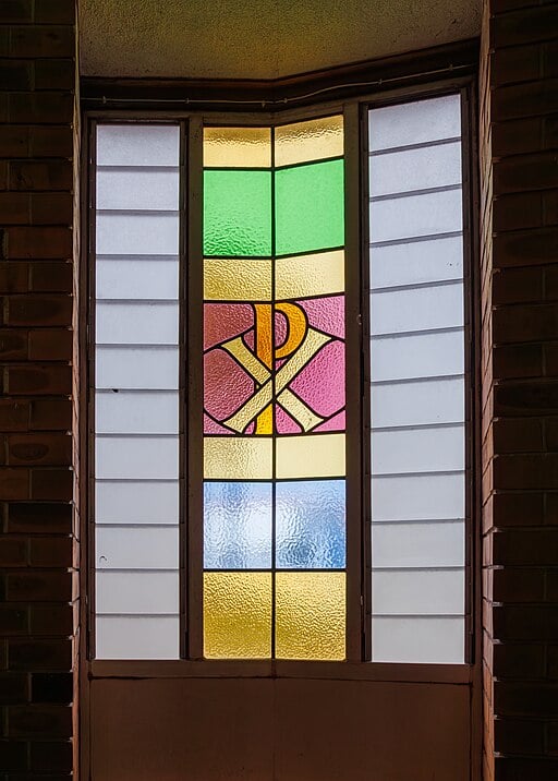 Stained glass window with multicoloured panels and a central chi rho symbol, flanked by frosted glass sections, set in a brick wall.