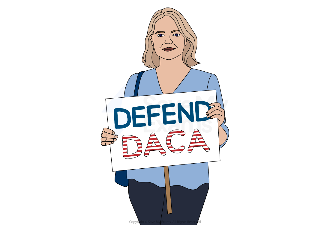 Person holding a sign reading "DEFEND DACA" during a protest. The sign features bold letters with an American flag pattern. Trees are visible in the background.