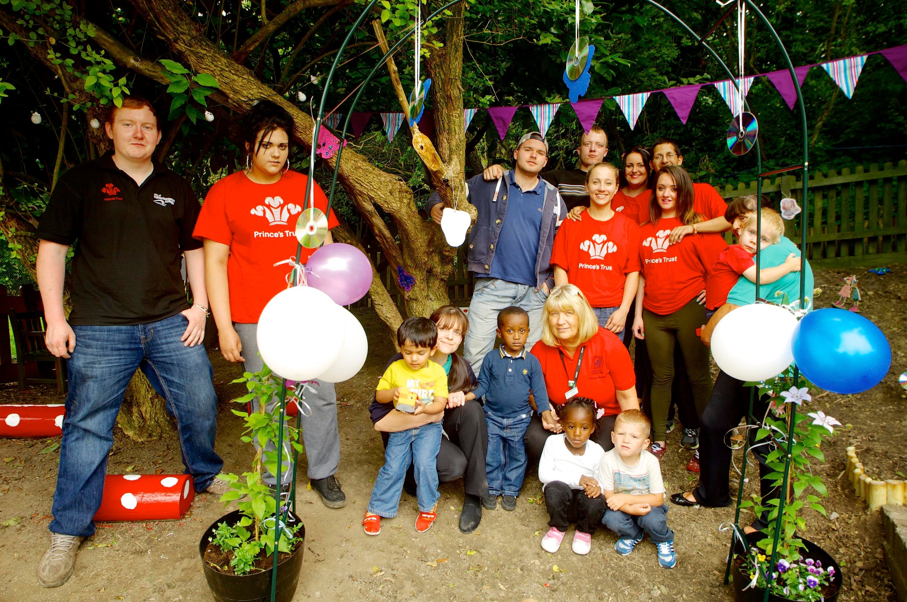 Group photo in a garden with adults in red shirts and children. Bunting, balloons, and plants decorate the scene. Forest background.