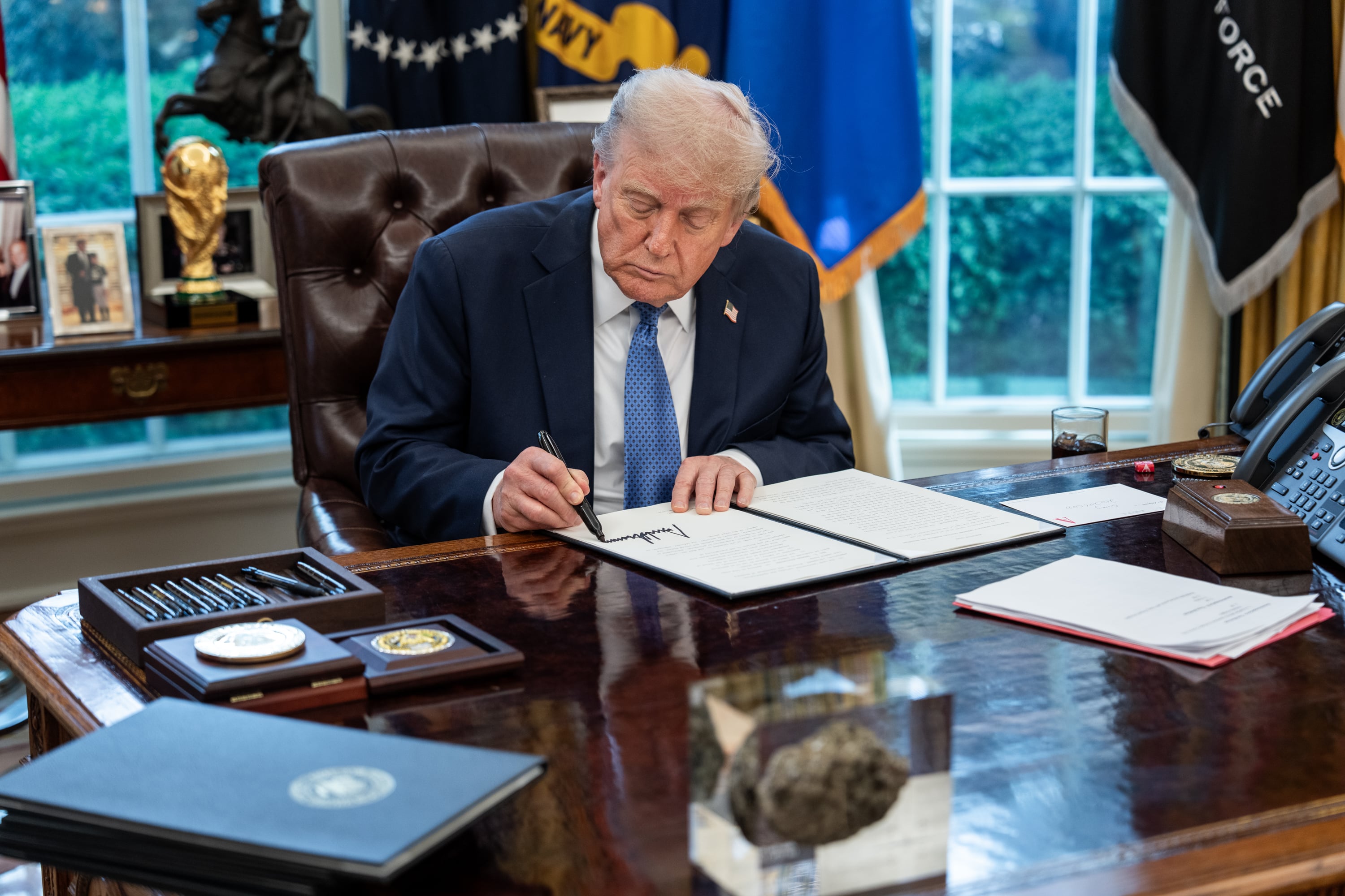 President Trump sits at the desk in the Oval office, signing a document