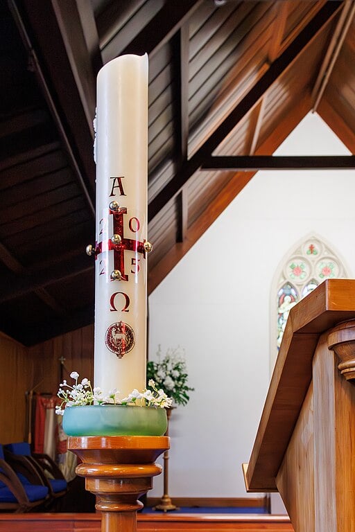A Paschal candle with religious symbols stands on a wooden holder in a church, near a lectern. Stained glass window visible in the background.
