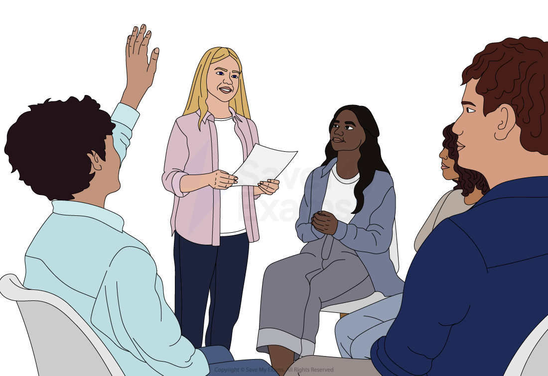 A blonde woman stands holding papers, speaking to a seated group. One person raises a hand, and others listen attentively, creating a discussion scene.