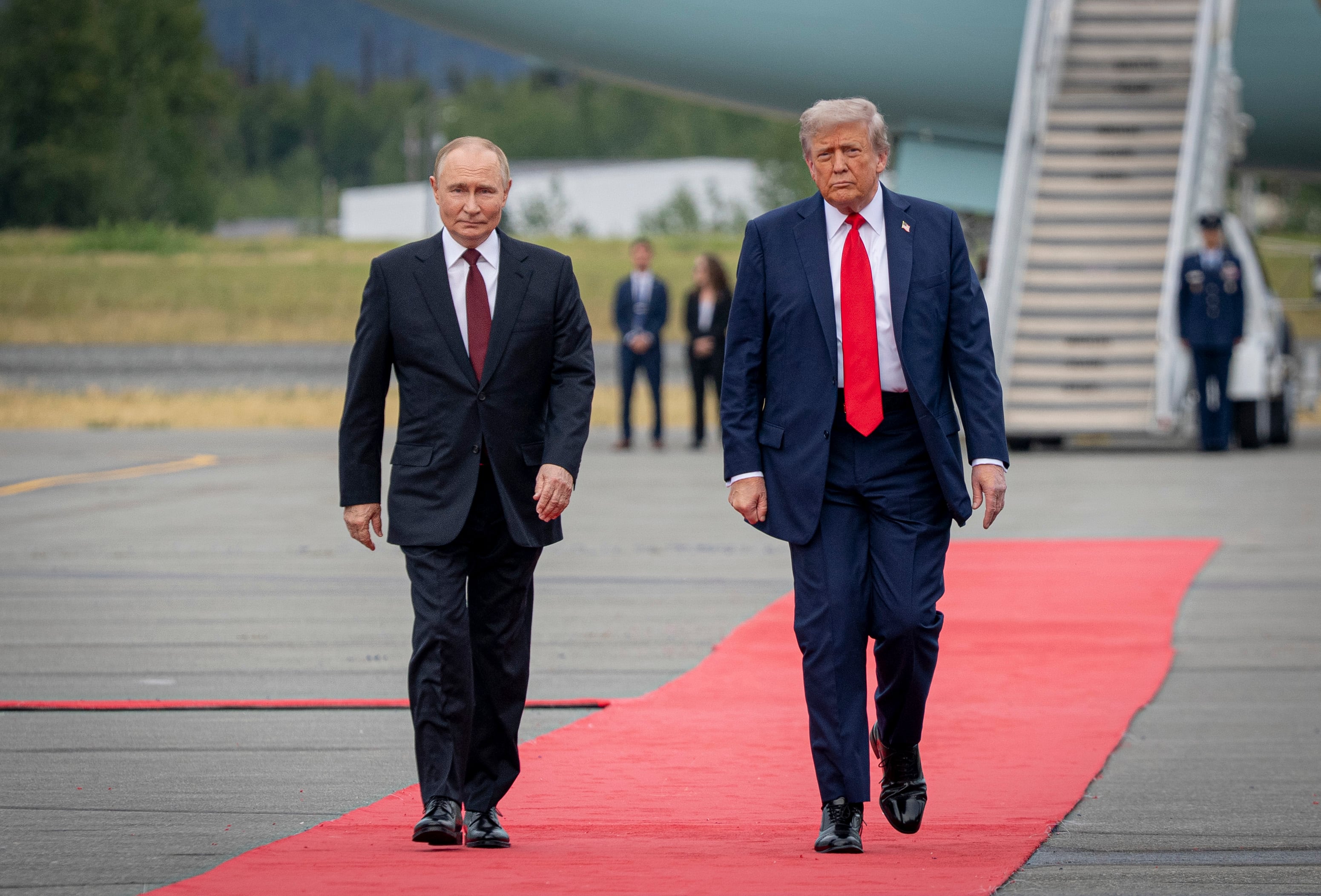 Two men in suits walk on a red carpet at an airport with a plane behind them and security personnel in the background.