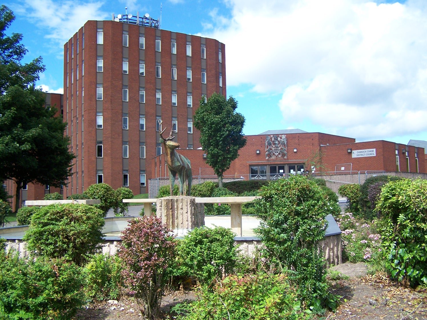 Red-brick council building with rectangular windows, a fountain featuring a deer statue, surrounded by landscaped gardens under a partly cloudy sky.