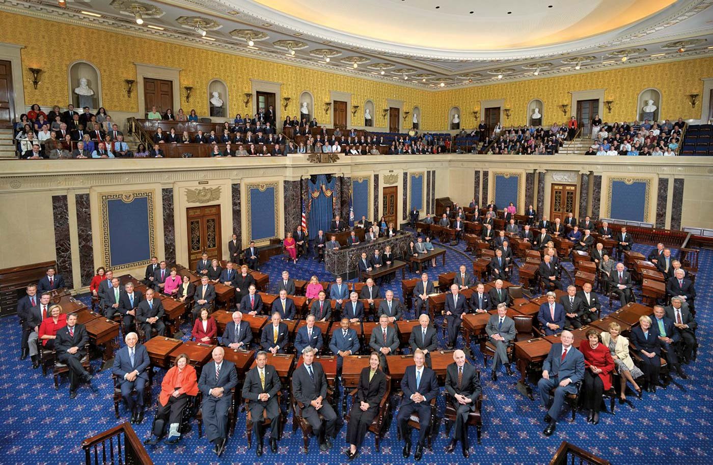 A large legislative chamber filled with suited individuals seated at desks, surrounded by an audience in elevated seating under a decorative ceiling.