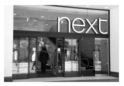 Entrance of a Next retail store with glass doors, large logo above, and reflections of people walking in a shopping area.