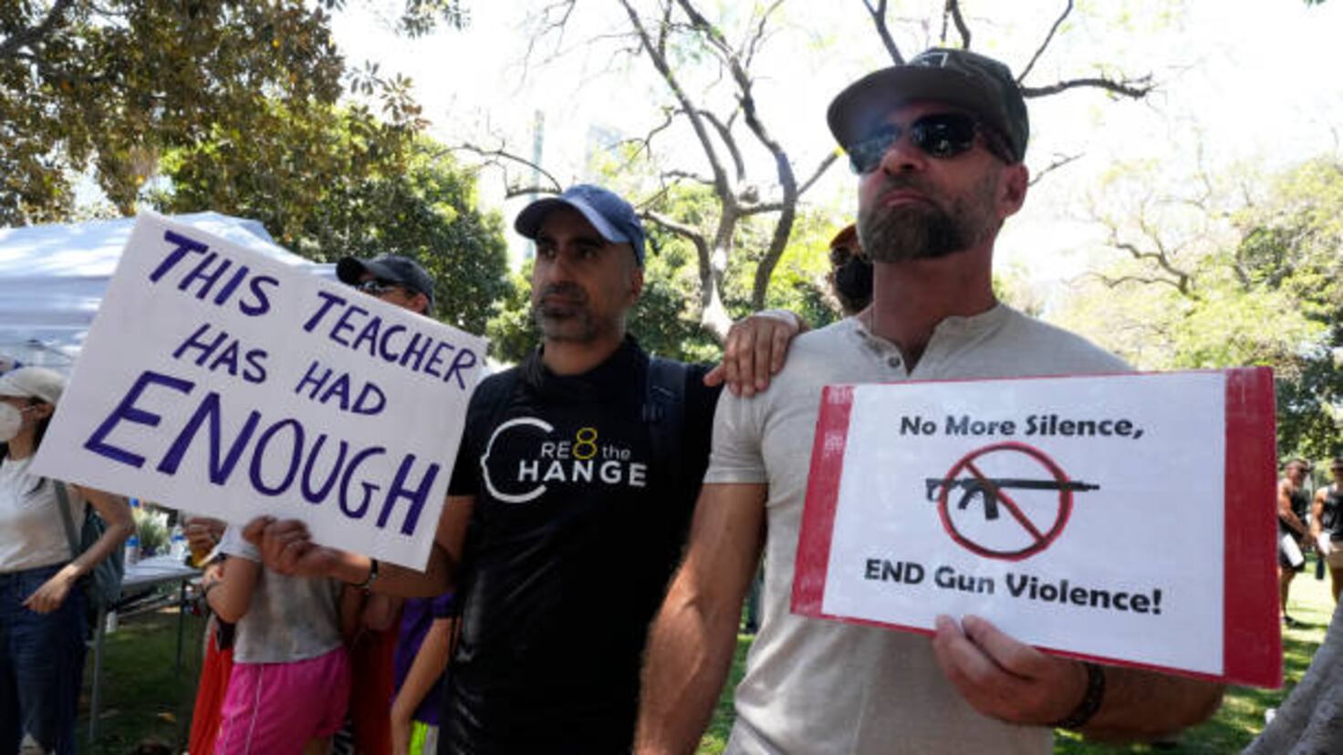 Protesters holding signs reading "This teacher has had enough" and "No more silence, end gun violence," stand outdoors on a sunny day.