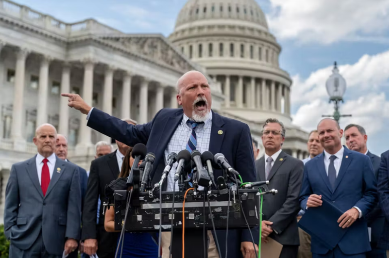 A group of men in suits stands in front of multiple microphones during a press conference outside a government building with a dome and columns.