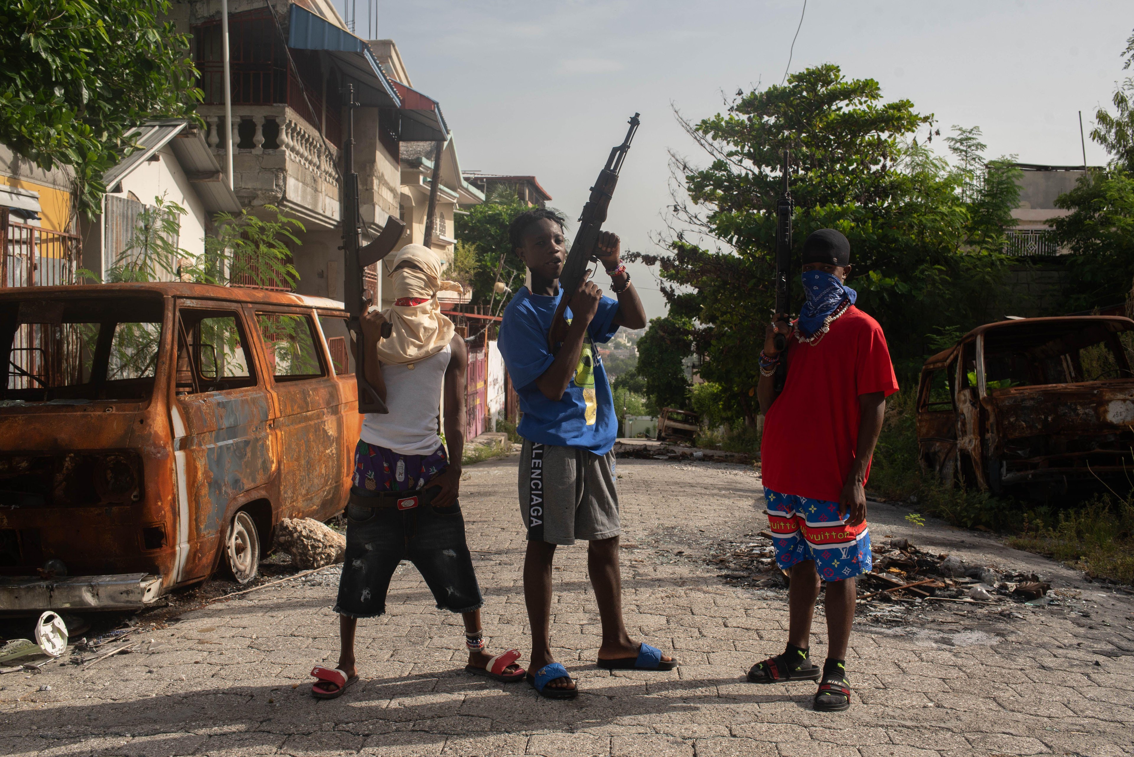 Young people, two masked and one armed with a gun, surrounded by a burnt-out vehicle