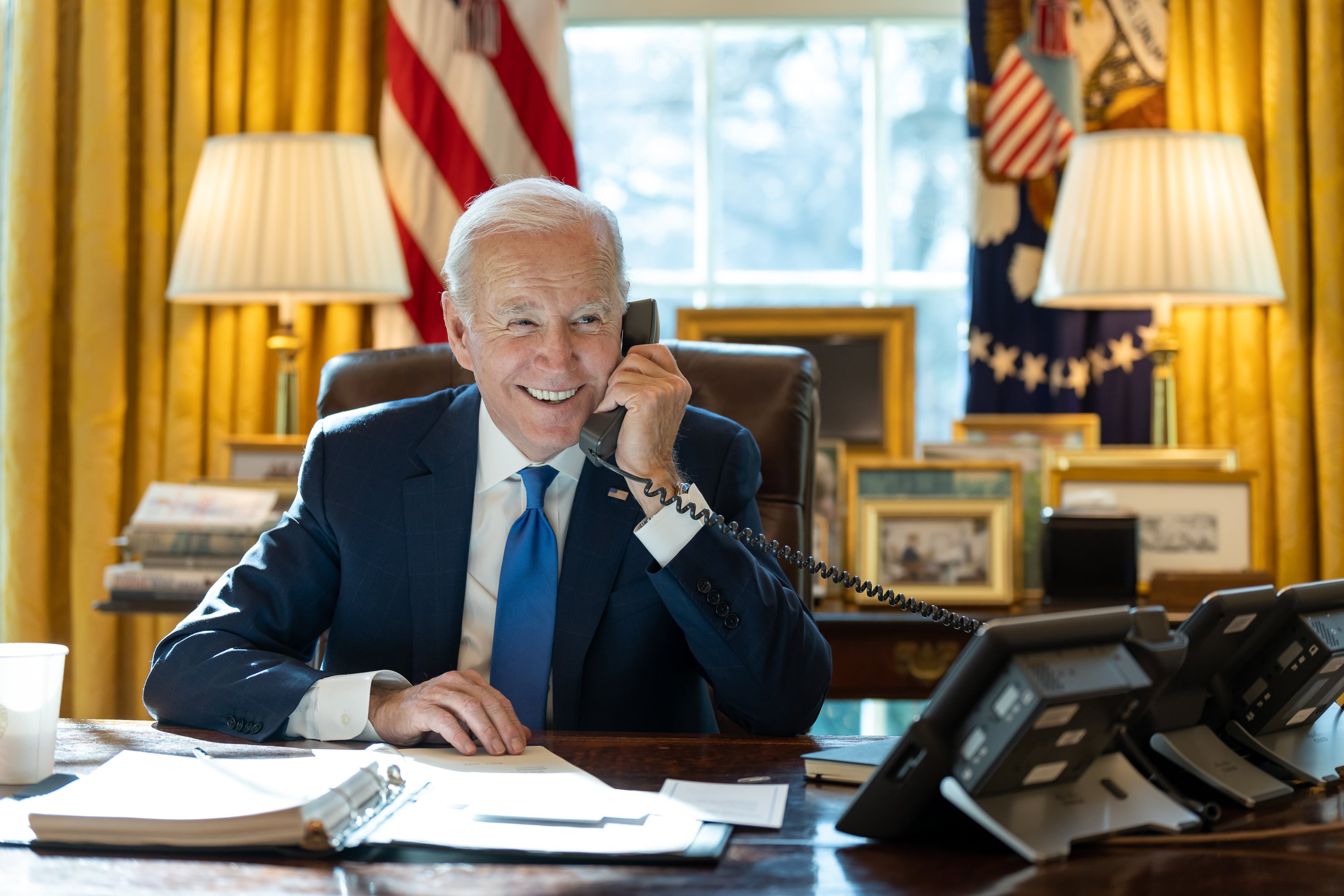 Smiling man in a suit speaks on the phone at a desk in an office with yellow curtains, framed photos, and flag decor.