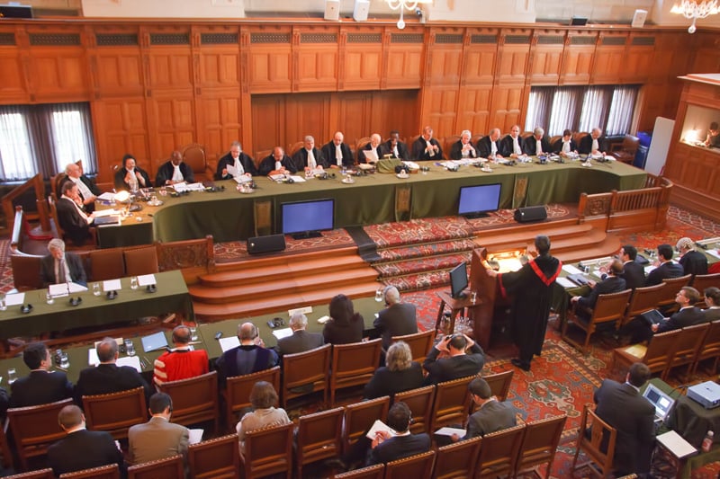 A formal courtroom with judges seated at a long table, lawyers presenting arguments, and an audience attentively listening in a wood-panelled room.
