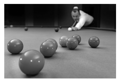 Pool player lining up a shot on a table with several balls scattered, focusing intently, in a black-and-white image.