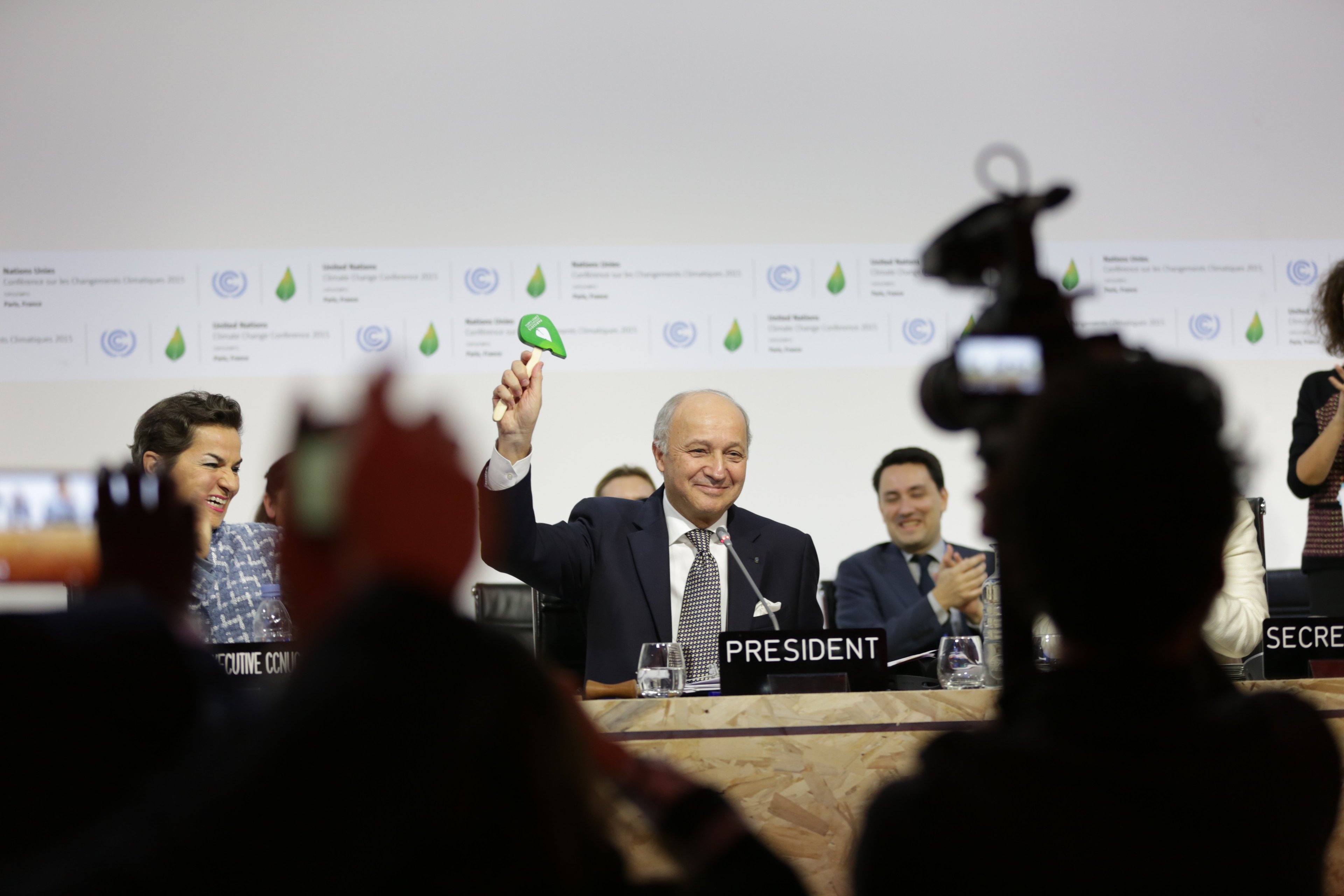 A man with a nameplate 'President' celebrates with other members of a group, with photographers in the foreground