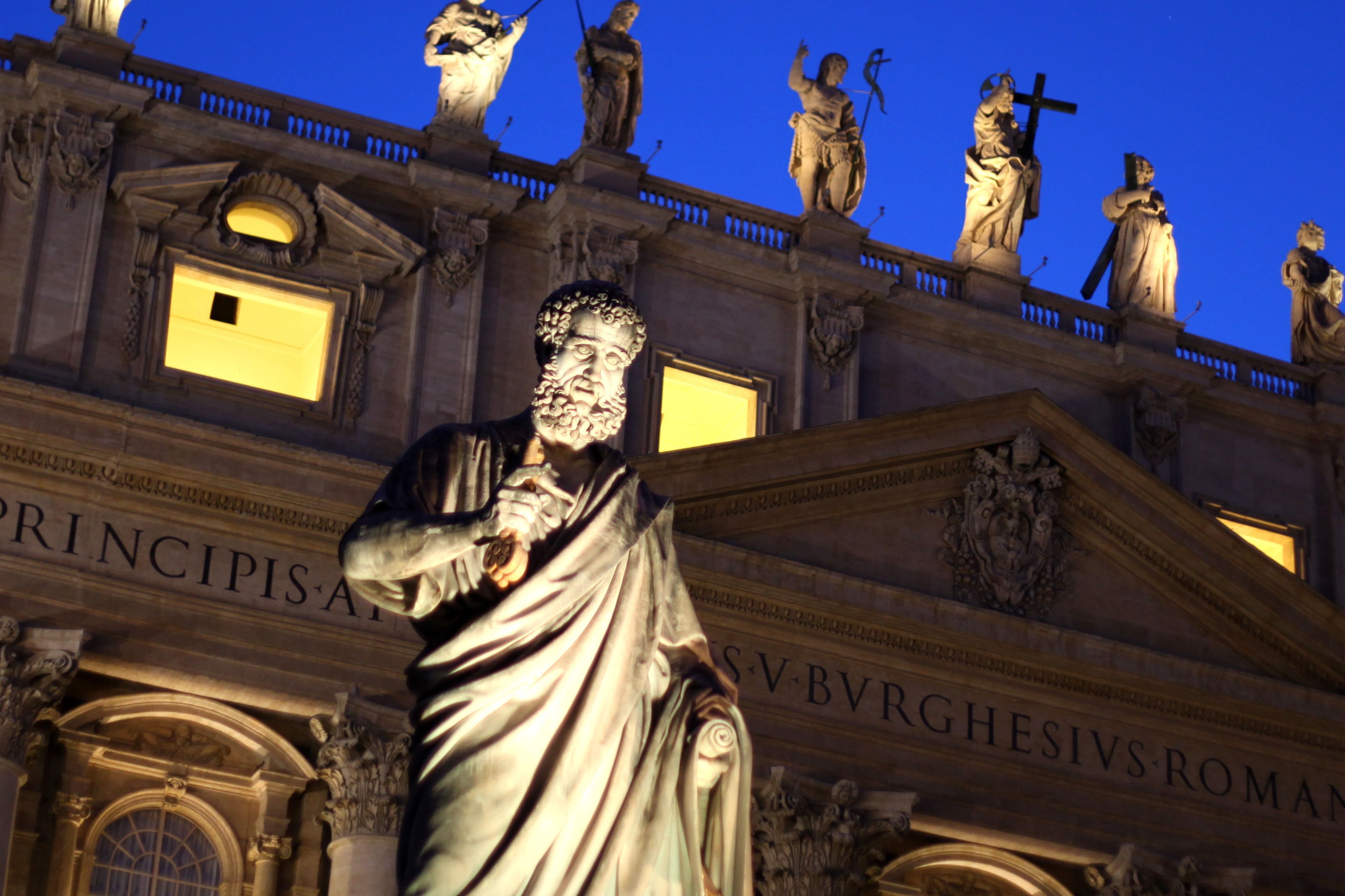 Illuminated statue of a bearded man holding keys in front of a lit classical building with statues against a twilight sky.
