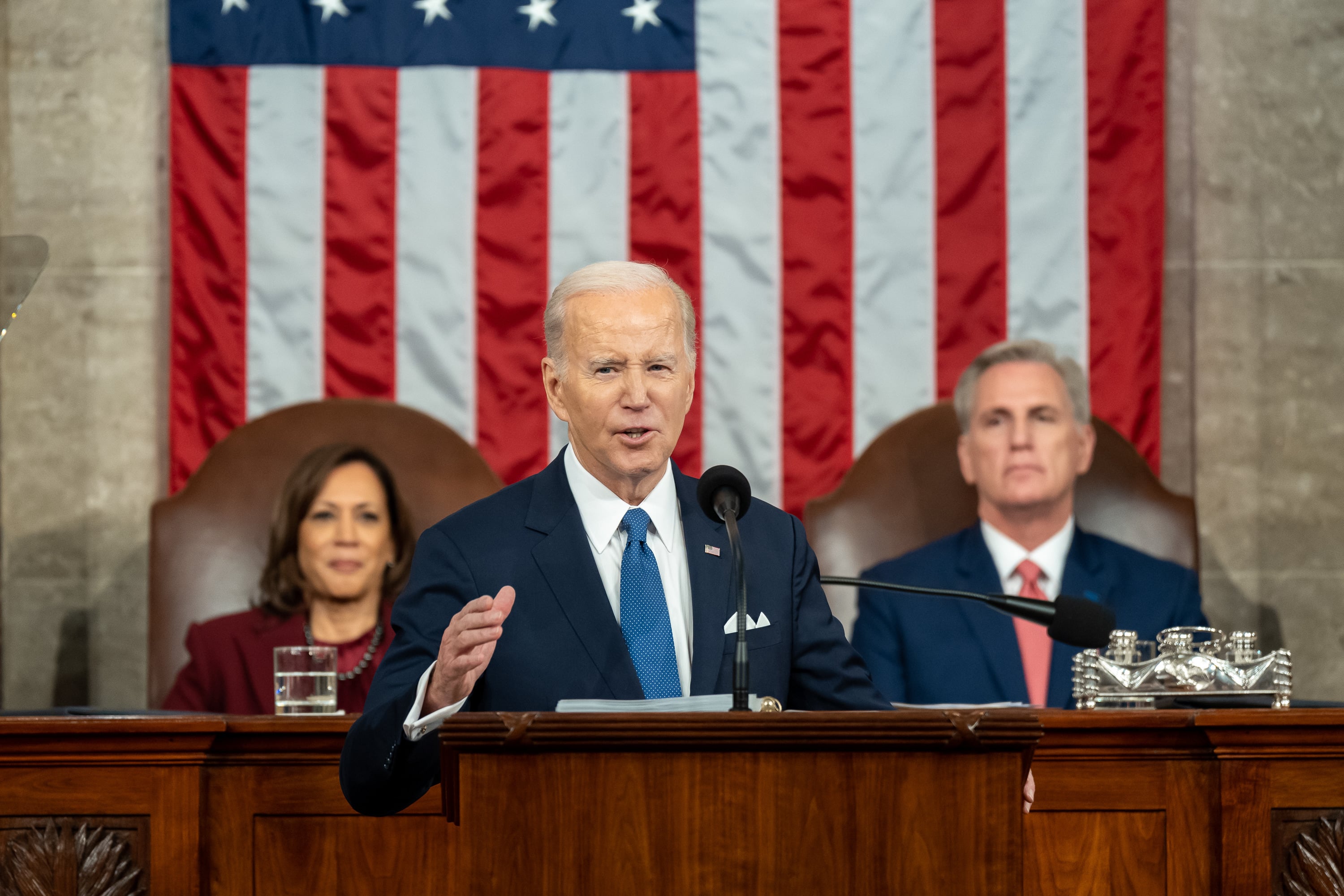 Man in a suit speaks at a podium, flanked by a woman and a man seated behind him, with an American flag in the background.