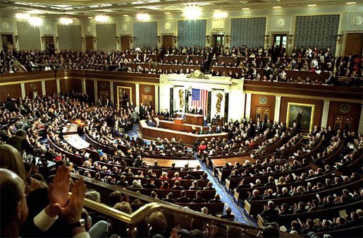 Large audience in government chamber with officials seated in semi-circle facing a podium, where a speaker stands, framed by an American flag.