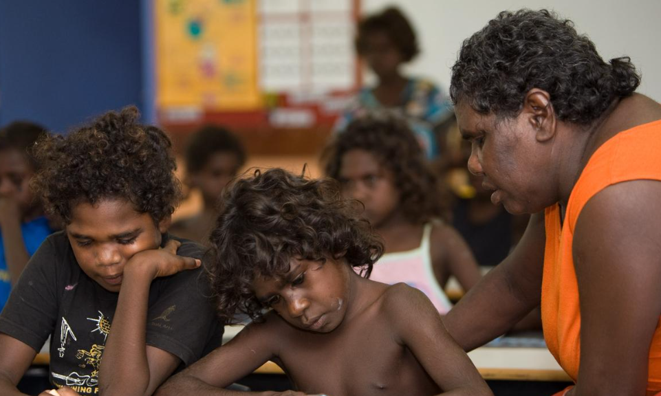 A teacher in an orange top assists two focused children with dark curly hair, in a classroom setting with more students in the blurred background.