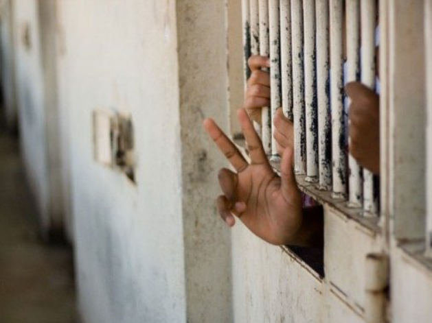 Hand making a peace sign through bars of a white, weathered prison cell, with another hand visible, suggesting confinement and hope.