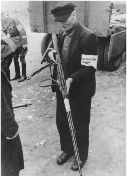 Elderly man in dark clothing holding a rifle, wearing an armband inscribed "Der Volkssturm," stands outdoors with a truck and Nazi soldier in the background.