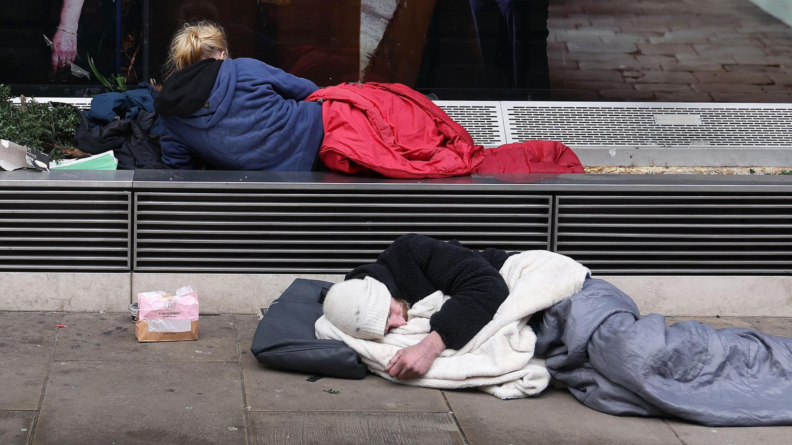 Two people sleeping outdoors on the pavement, one under a red blanket on a bench, the other under a white blanket on the ground.