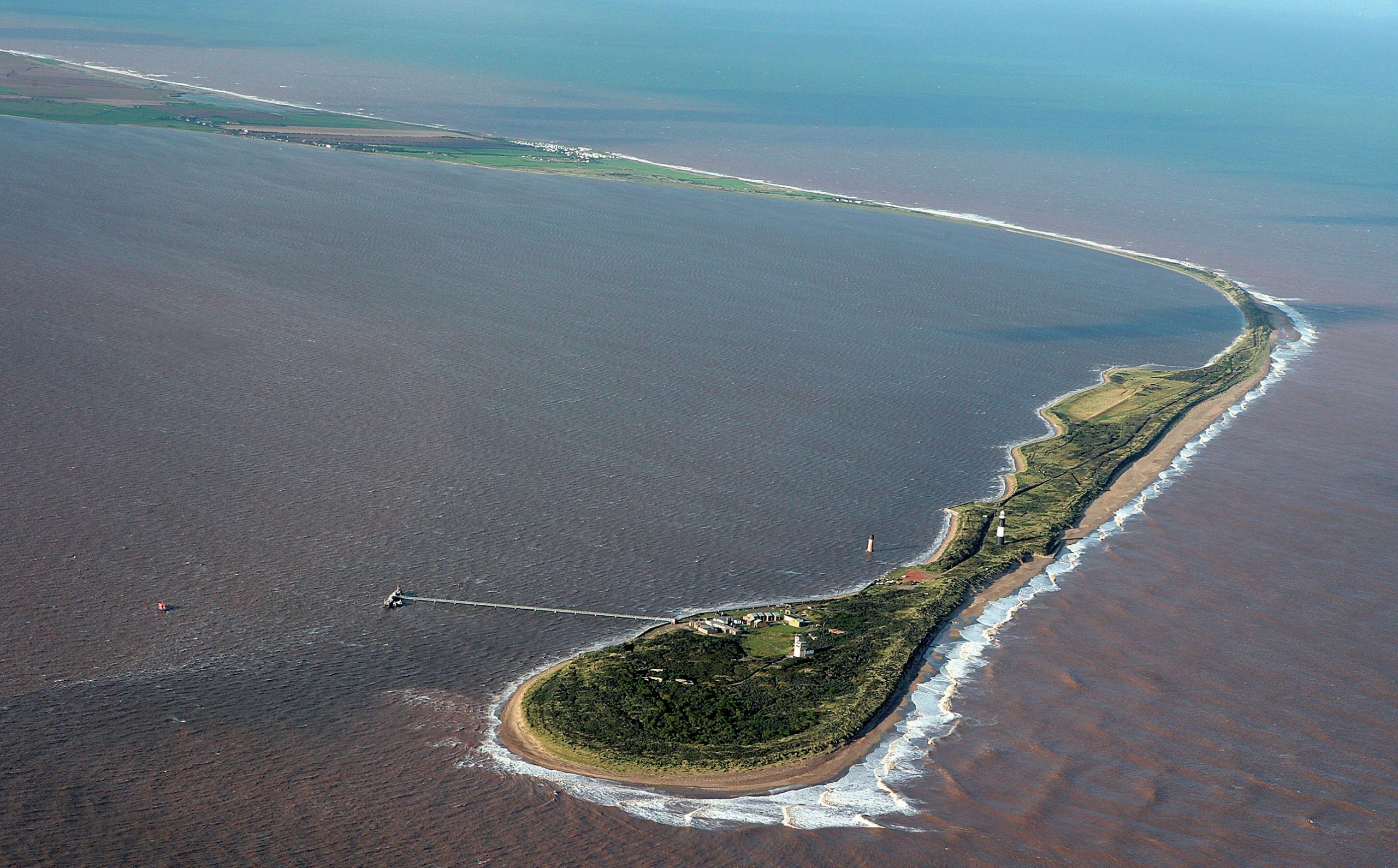 Aerial view of a narrow landmass with lush greenery and a few structures, surrounded by brownish sea waves, connected to the mainland by a thin strip.